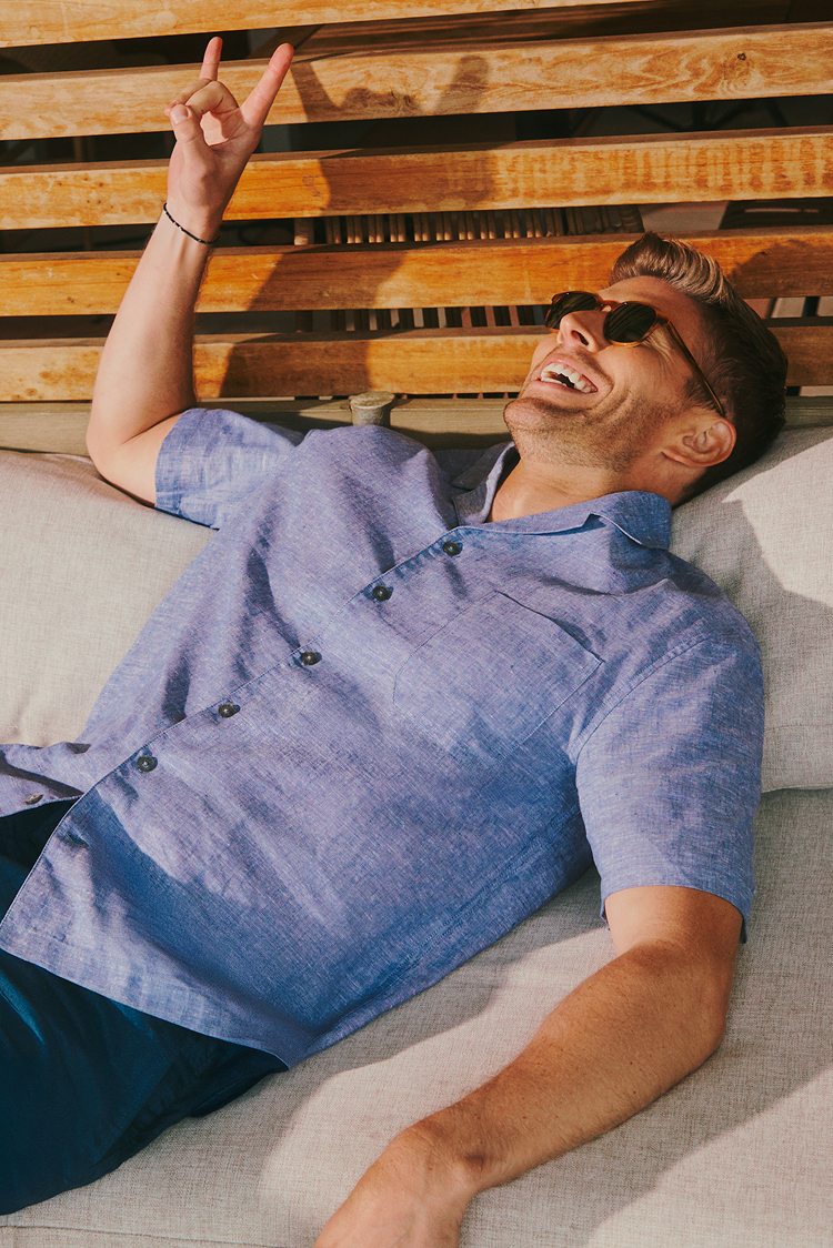 Man in a blue shirt and sunglasses lying on a couch, making a peace sign.