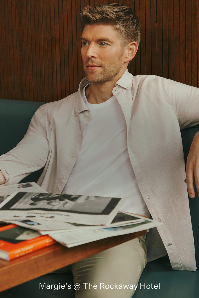 Man sitting on a couch with books and magazines, wearing a light-colored shirt over a white t-shirt.